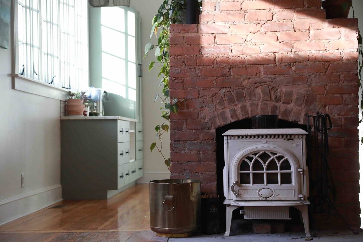 Interior of a cozy room with a white vintage stove against a red brick fireplace, surrounded by green plants and natural light from large windows.
