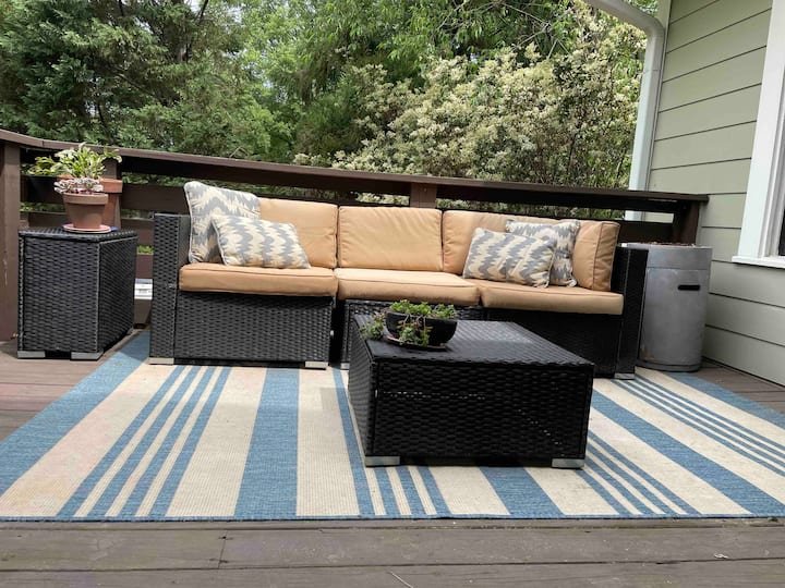 Outdoor patio with a wicker sofa set, tan cushions, striped rug, and potted plants, surrounded by greenery.
