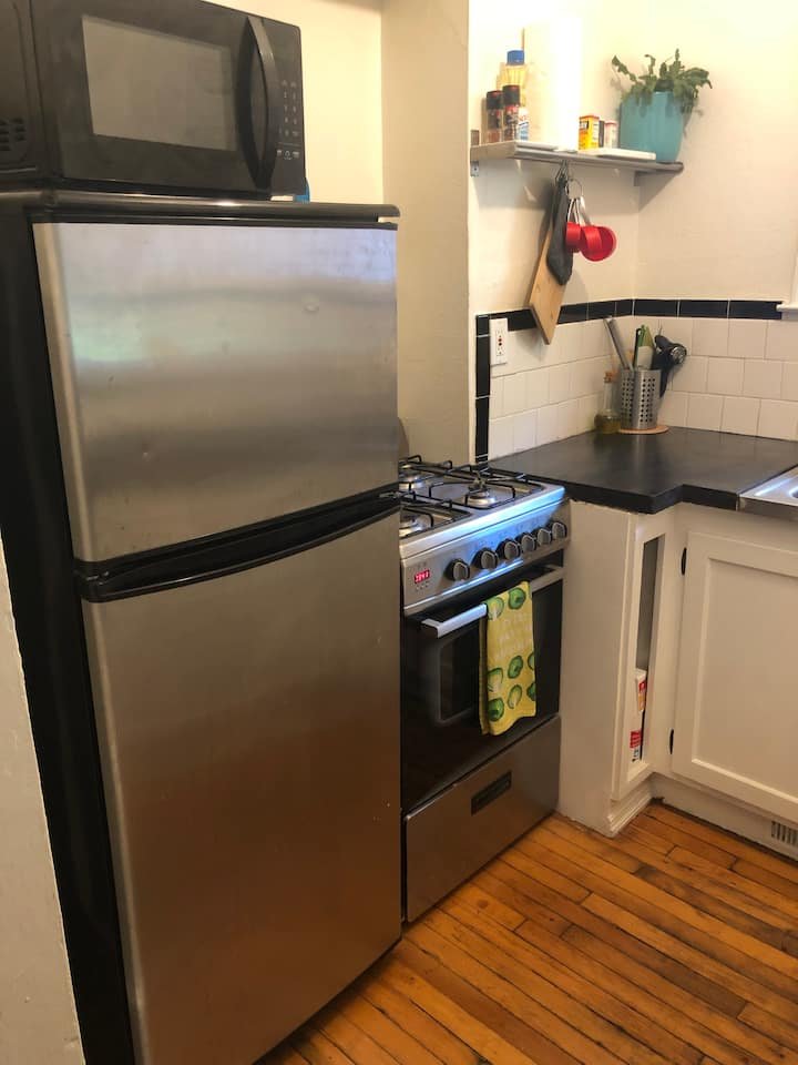 Small kitchen corner with a stainless steel refrigerator, black microwave on top, gas stove with an oven, wooden cutting board on the wall, a green towel hanging on the oven door, white cabinets, and wooden flooring.