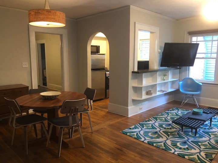 Cozy living room with round wooden dining table, chairs, and light fixture. A modern area rug and TV, with a blue chair by the window, and an archway leading to the kitchen are visible.