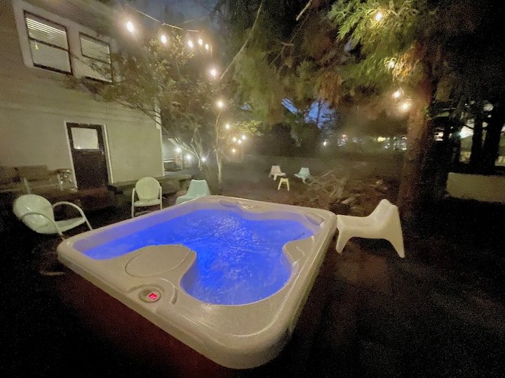 An outdoor hot tub illuminated with blue lights at night, surrounded by outdoor chairs, string lights, and trees.