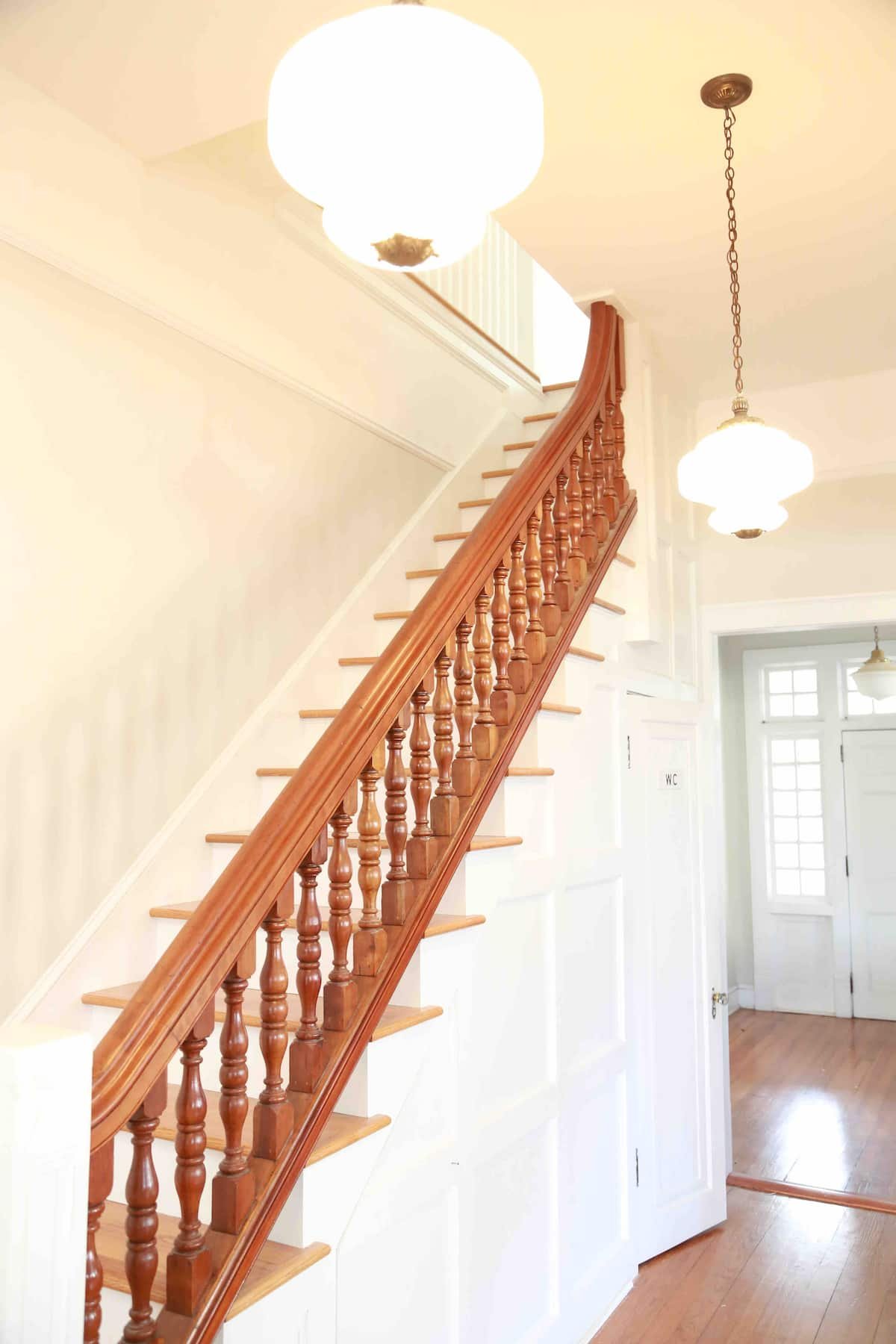 Wooden staircase with ornate banister in a bright interior space, hanging pendant lights, and a door labeled WC near large windows.
