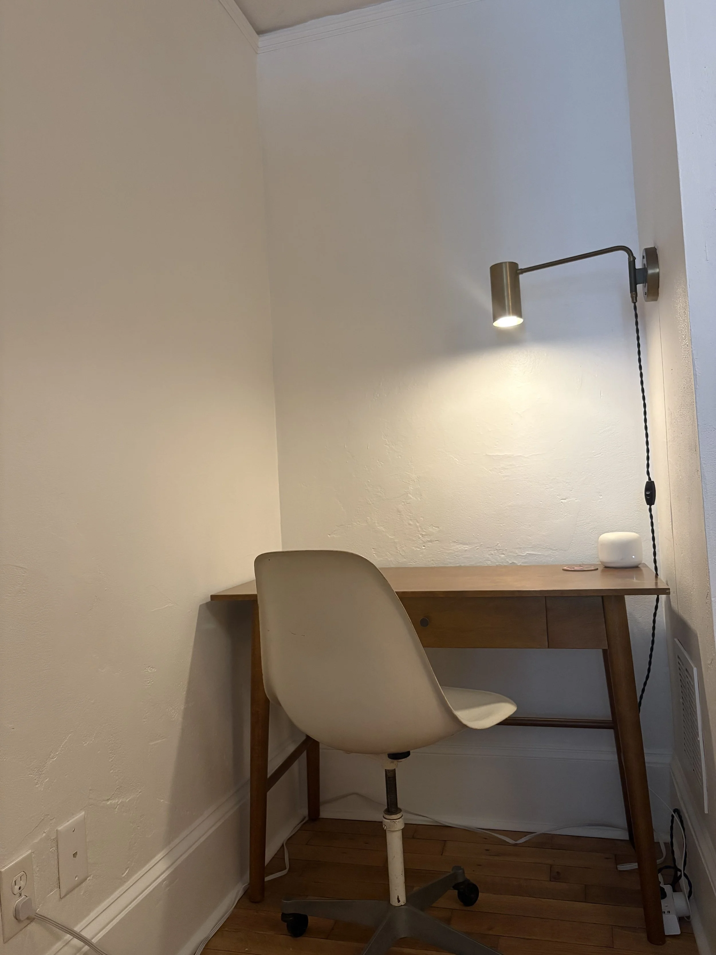 A small writing desk with a white swivel chair in front of it, illuminated by a wall-mounted adjustable lamp, in a corner of a room with hardwood flooring and white walls.