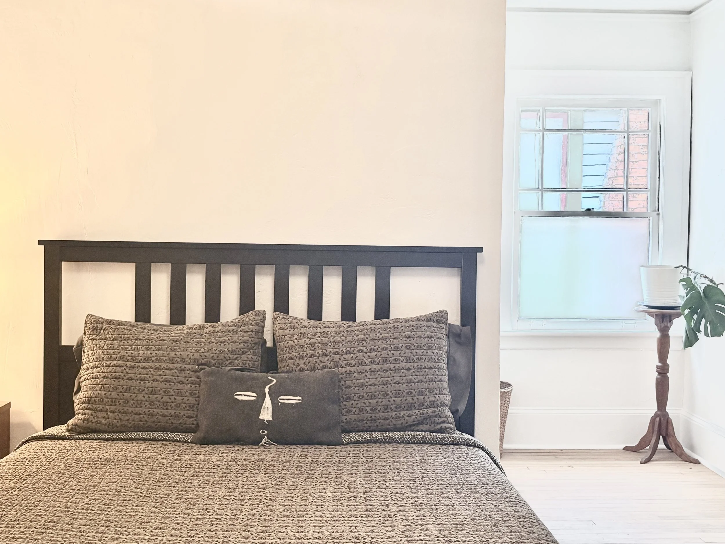 A bedroom with a black slatted headboard, two large patterned pillows, a decorative pillow with a face design, a window with blinds, a wooden side table with a plant in a white pot, and light-colored walls and flooring.