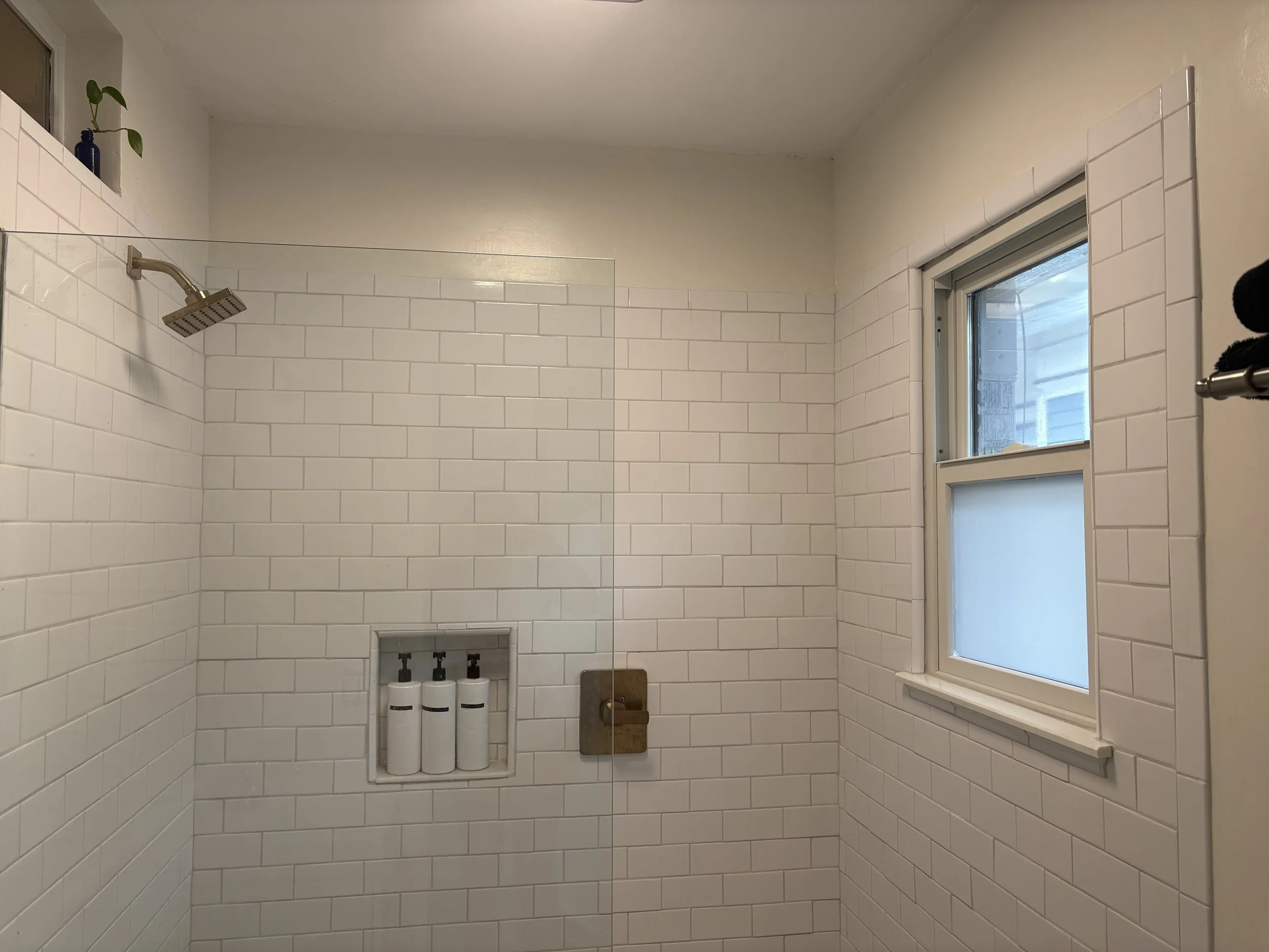 A shower area with white subway tile walls, a window with frosted glass, and a built-in shelf with three bottles of shower products. The shower has a brass shower fixture and a glass partition.