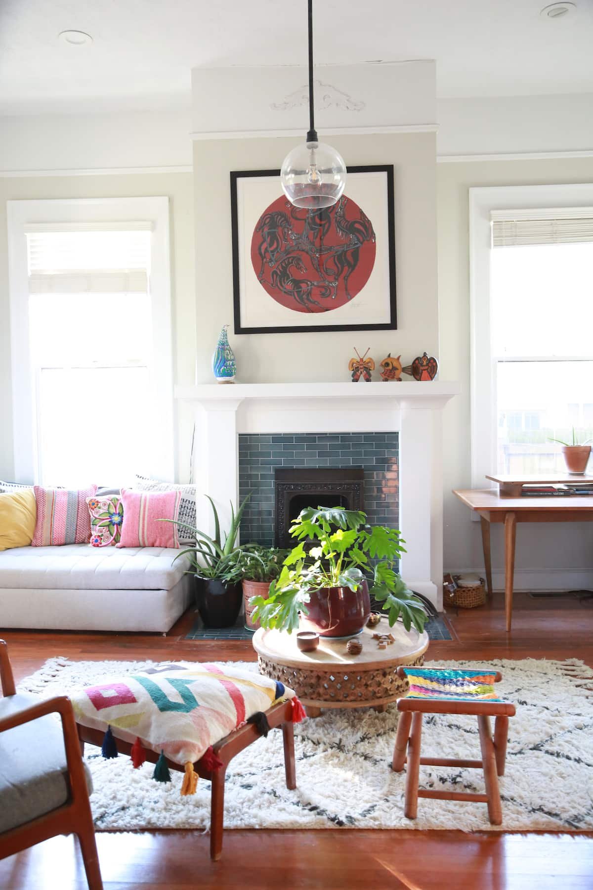 Living room with white sofa, plants, a fireplace, colorful pillows, a wooden coffee table with decorative items, and framed artwork above the fireplace.