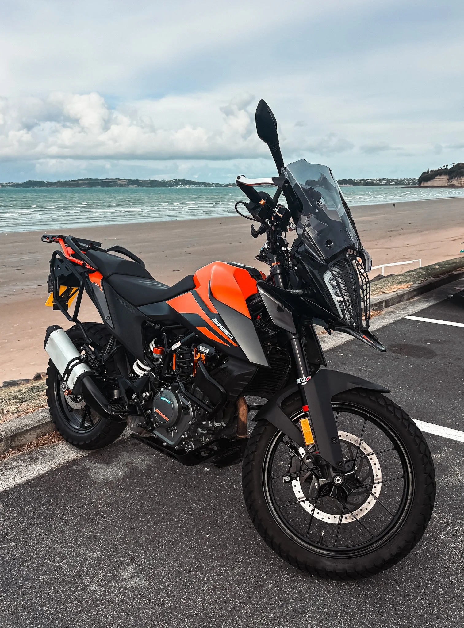 Orange and black motorcycle parked near a beach with ocean and cloudy sky in the background.