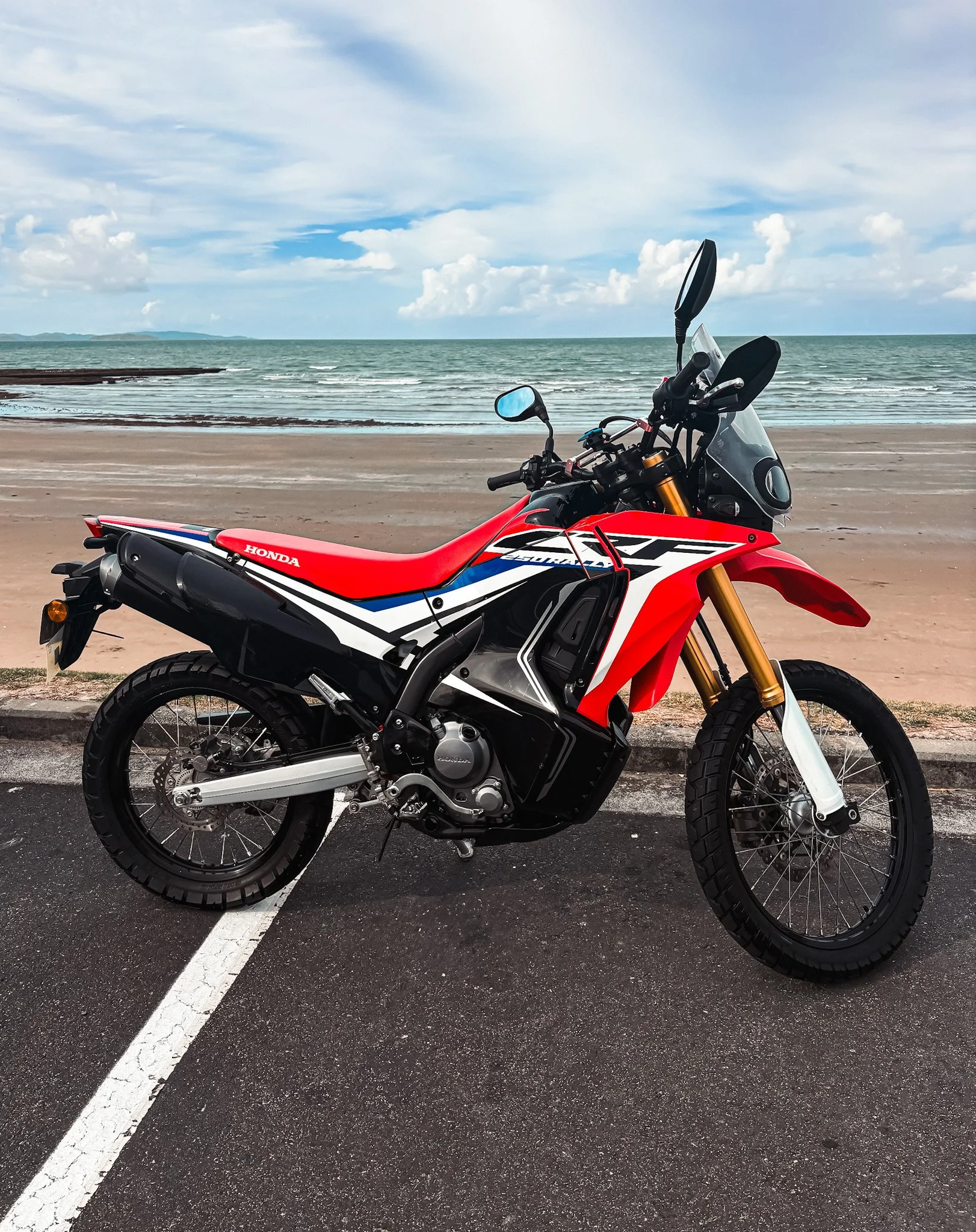 Red and white motorcycle parked on a beachfront road with ocean in the background.