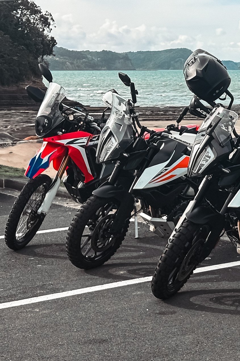 Three parked adventure motorcycles overlooking a scenic coastal view with mountains and sea in the background.