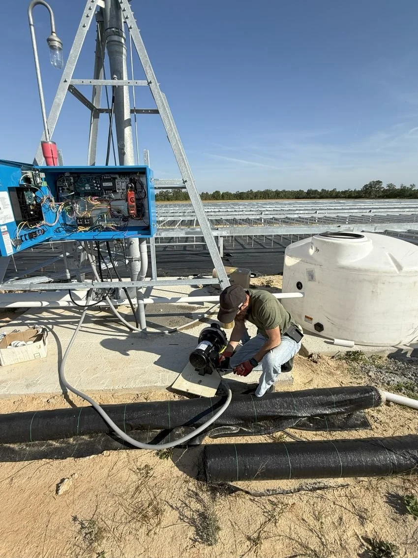 Technician working on electrical equipment outdoors on a farm, with solar panels and water storage tank in the background.