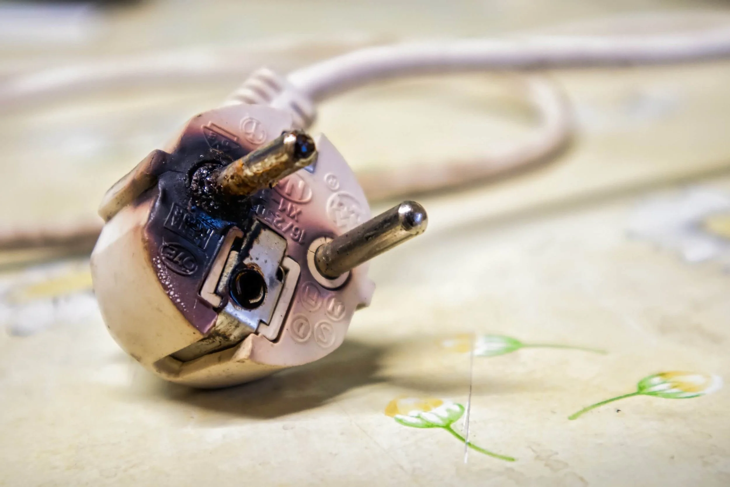 A close-up of a damaged electrical plug with burnt marks and exposed metal prongs resting on a light-colored surface with flower designs.
