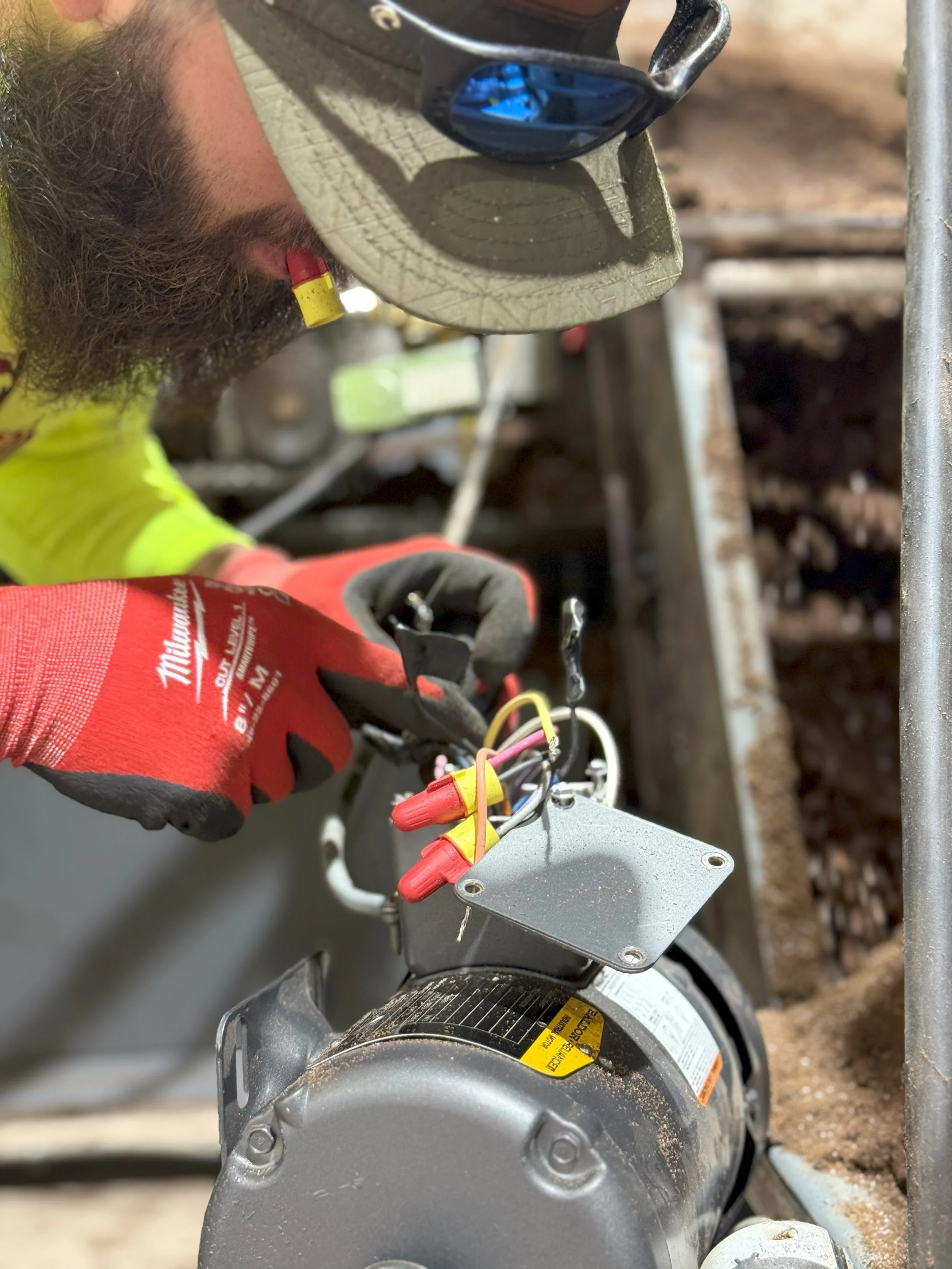 A worker wearing a gray cap, sunglasses, and red gloves working on electrical wires connected to a motor or pump, with dirt and metal frame nearby.