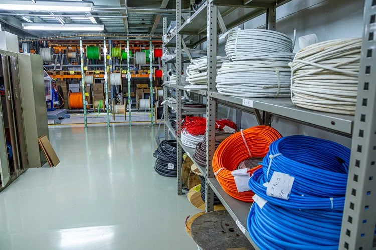 Industrial storage room with shelves containing coils of white, red, black, orange, and blue cables, and spools of wire in the background.