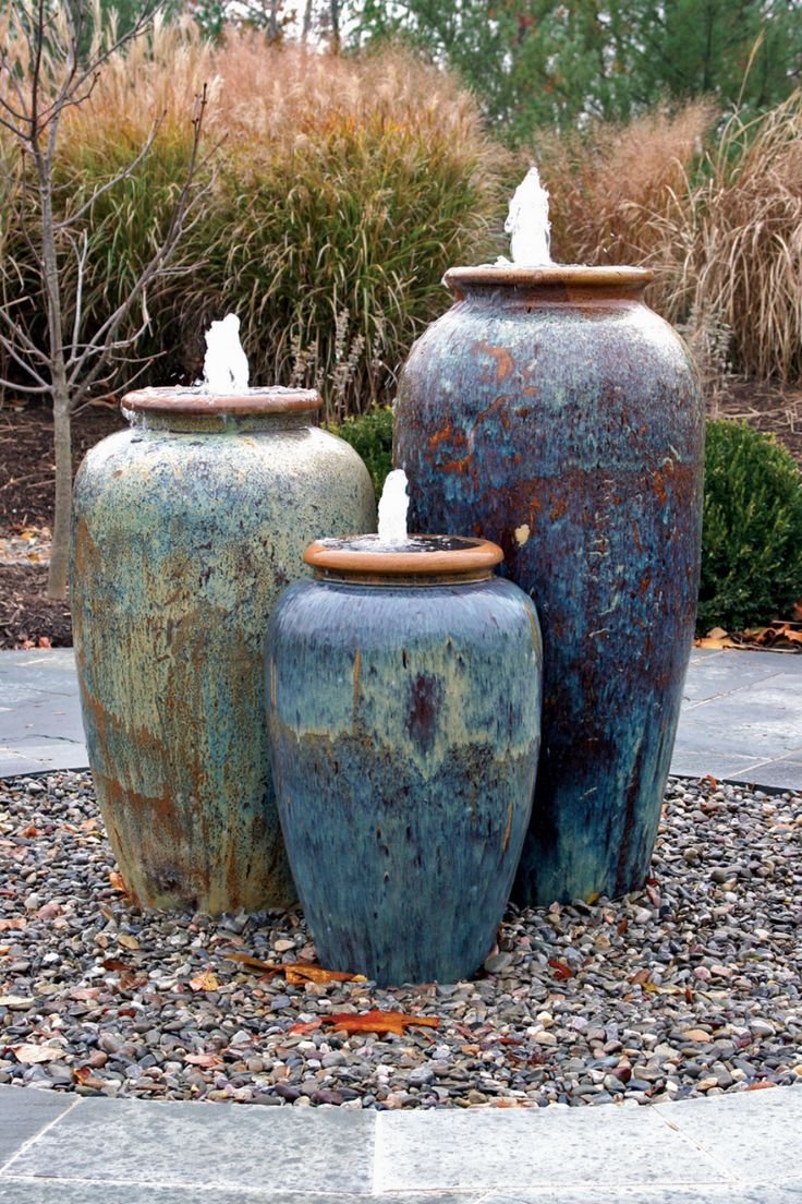 Three ceramic vase fountains with water flowing from the top, set on a bed of pebbles in a garden area with tall grasses in the background.