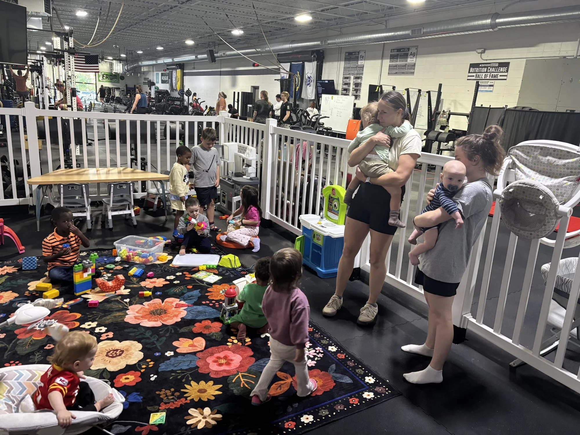 Toddlers playing on a flower rug in the fenced kids area while two moms hold babies, with the gym floor behind
