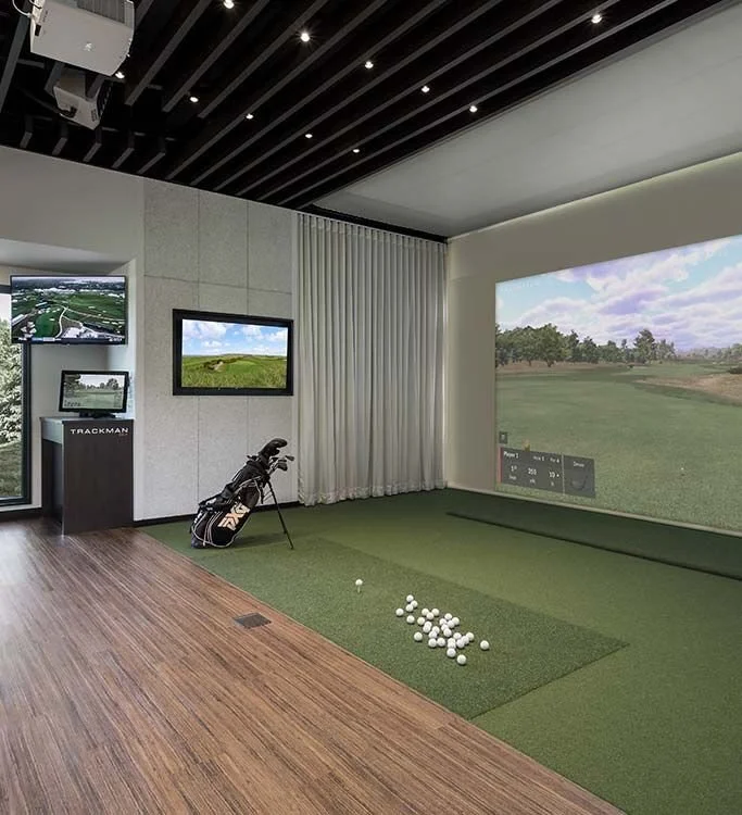 Indoor golf simulation room with a large screen displaying a golf course, a golf club with balls on a green mat, and multiple monitors showing golf scenery.