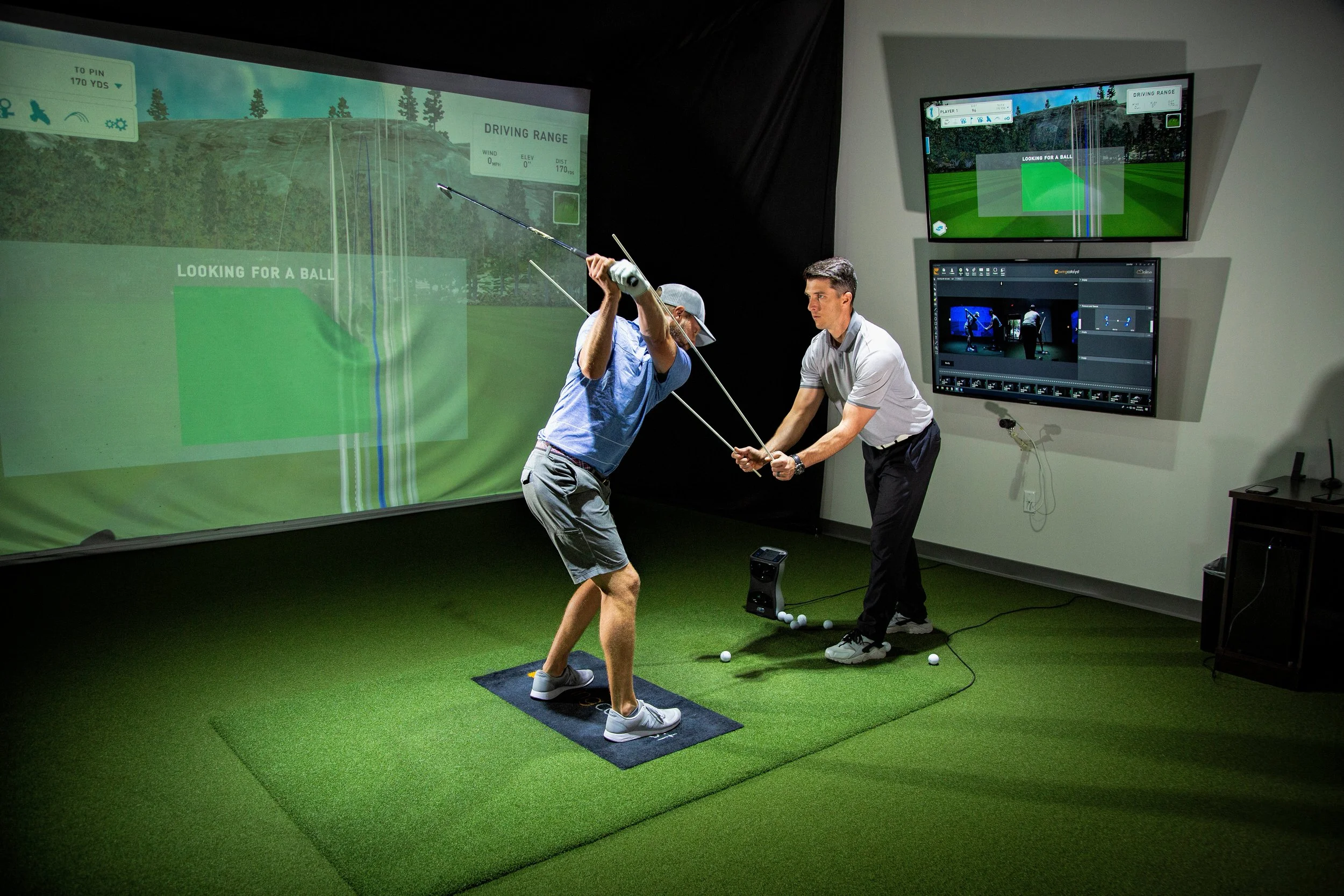 Indoor golf simulation with two men, one swinging a golf club and the other assisting, on artificial turf with multiple screens displaying golf courses.