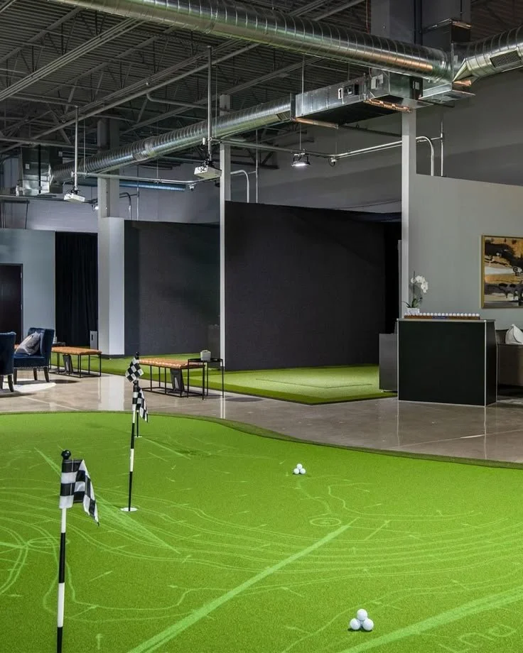 Indoor golf practice area with artificial turf, golf balls, and flags, in a modern room with seating and artwork.