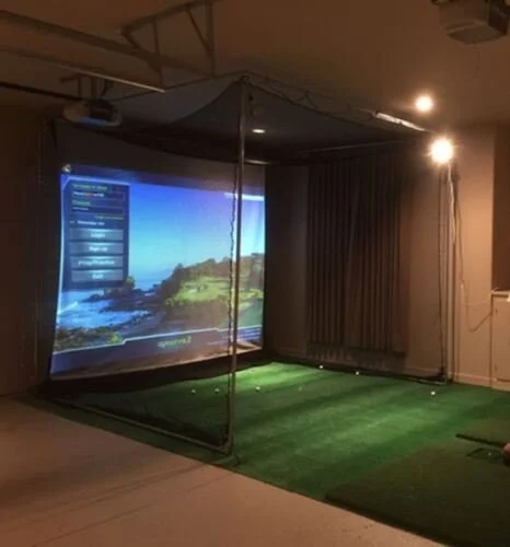 Indoor golf simulator with a projected screen showing a scenic coastal view, with a green artificial turf and a net for swing practice.
