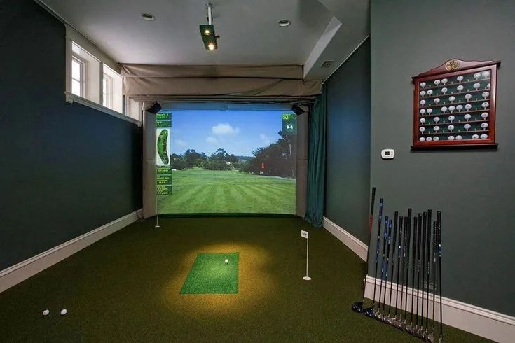 Indoor golf simulator room with a screen displaying a golf course, golf clubs lined against the wall, and a golf ball on a small artificial green mat.