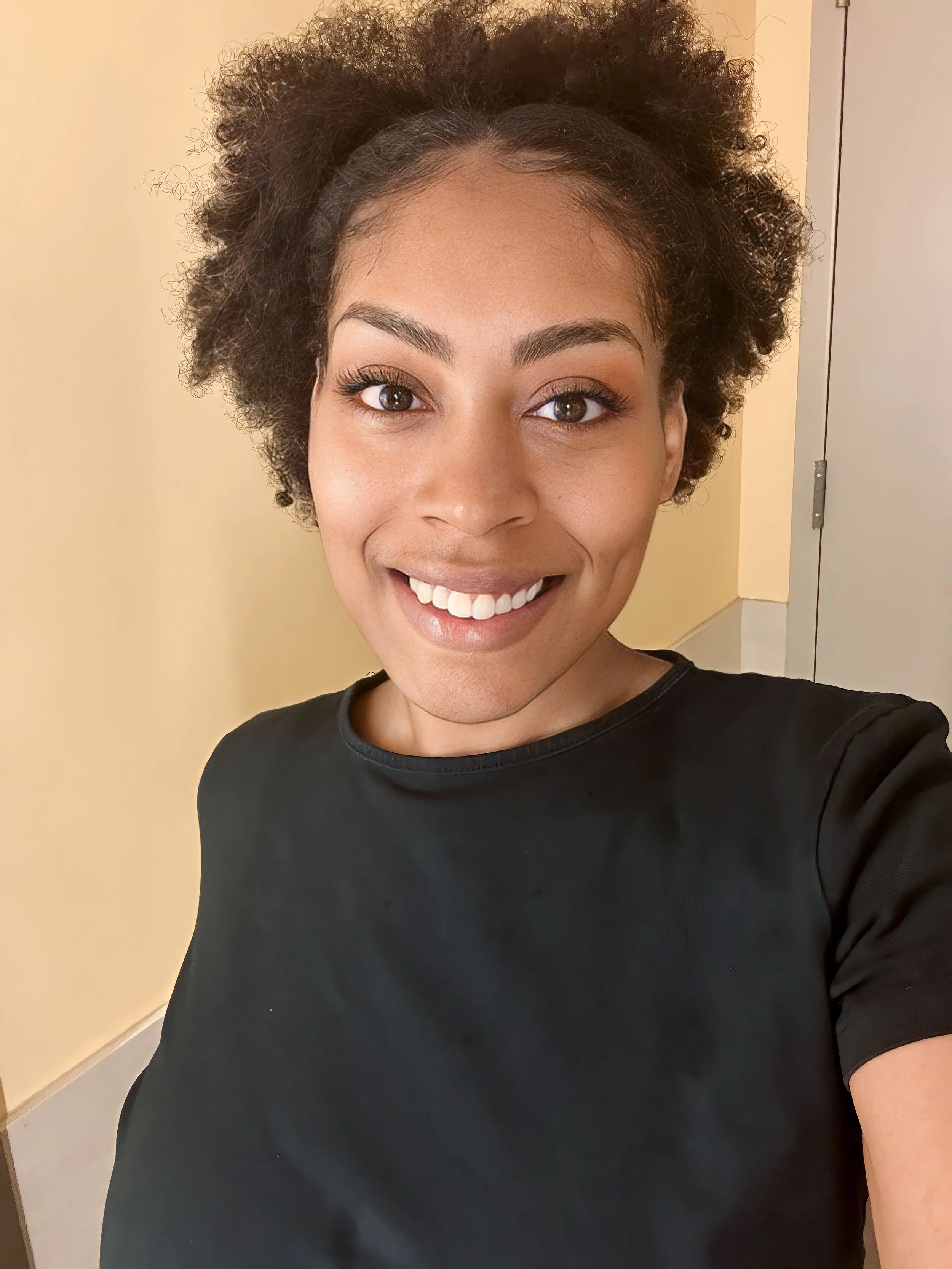 A young woman with curly hair smiling and taking a selfie in a room with a beige wall and a door in the background.