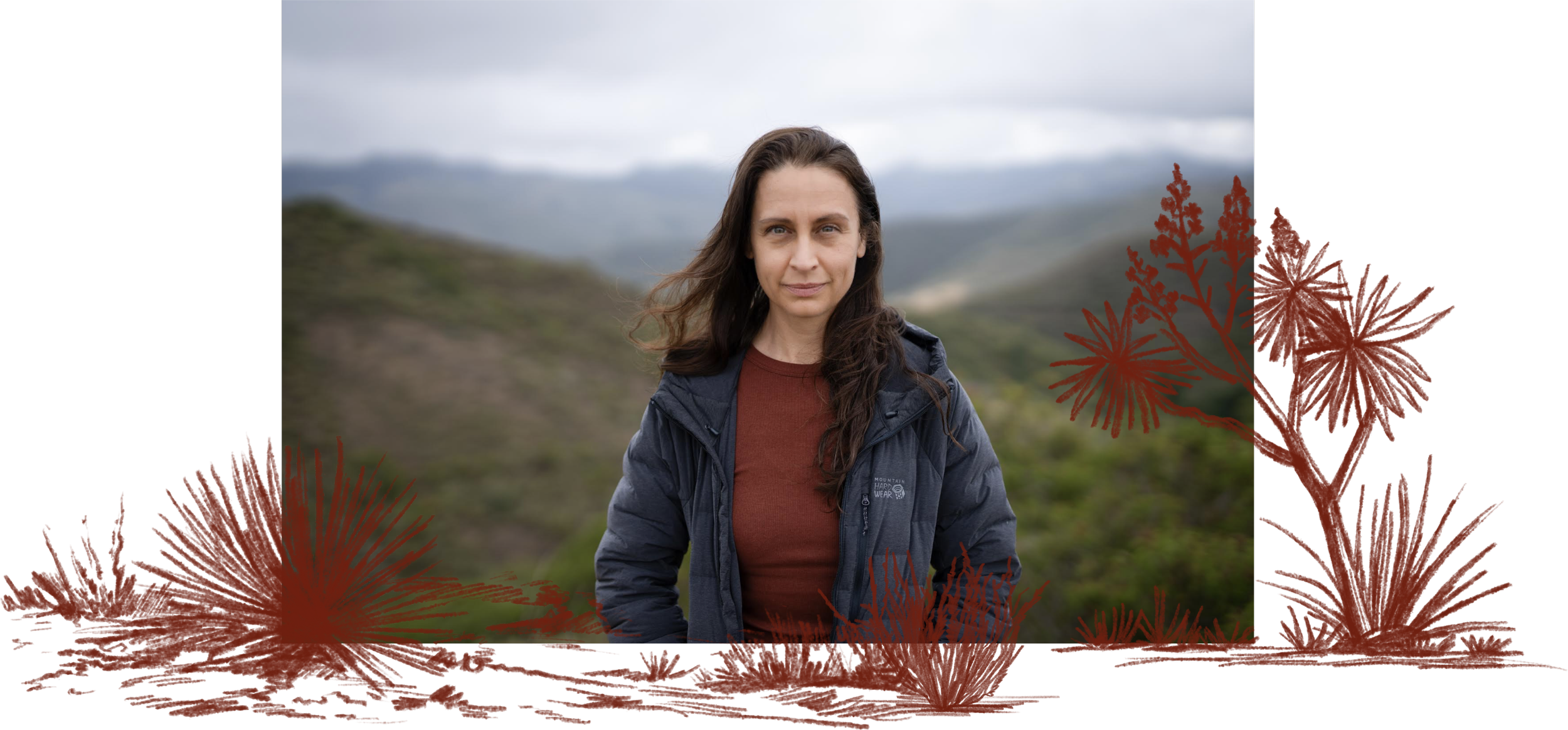 A woman standing outdoors in a natural landscape with hills and cloudy skies, wearing a blue jacket and a maroon shirt.
