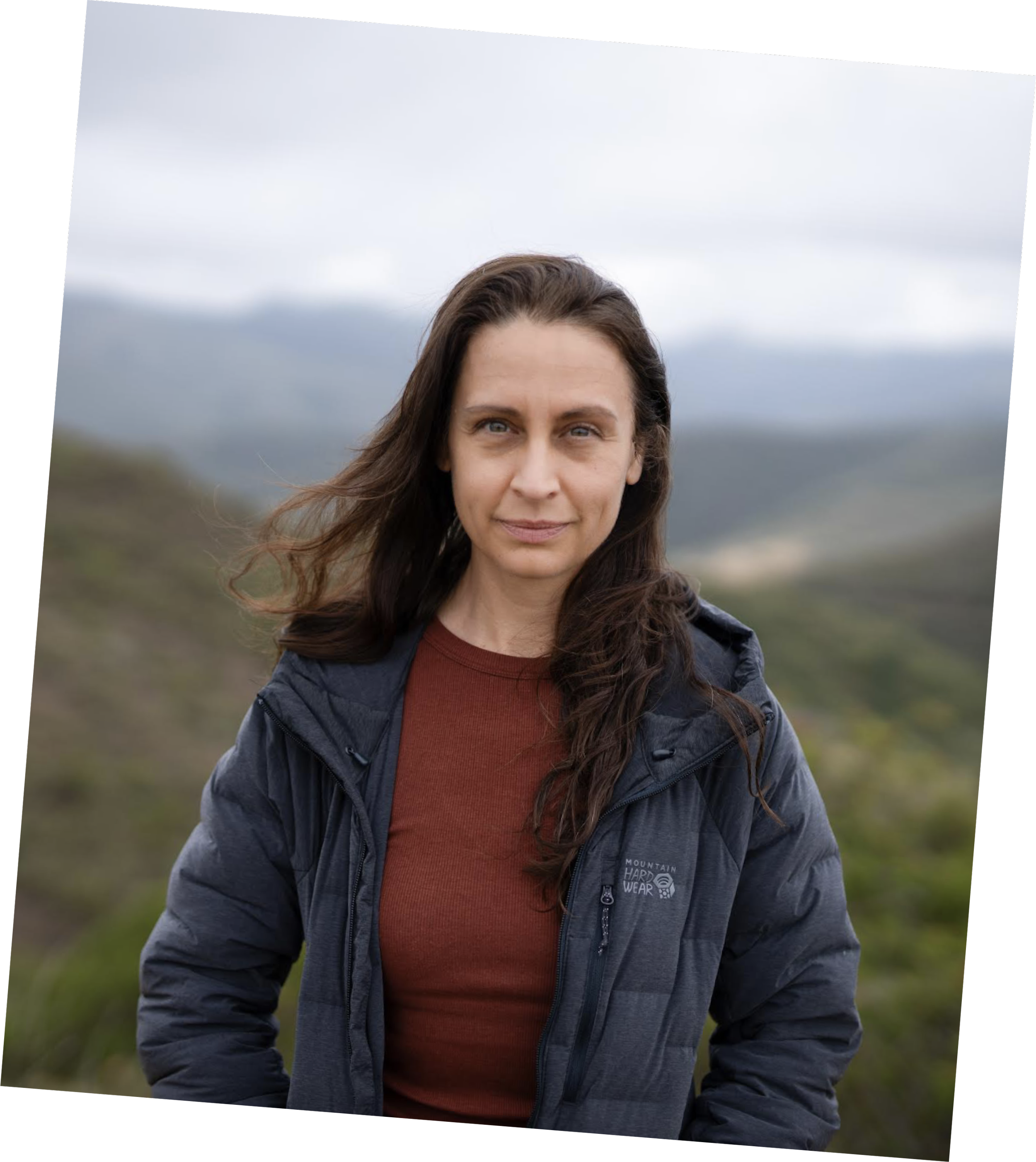A woman with long dark hair standing outdoors on a cloudy day with mountains in the background, wearing a blue jacket and a brown shirt.