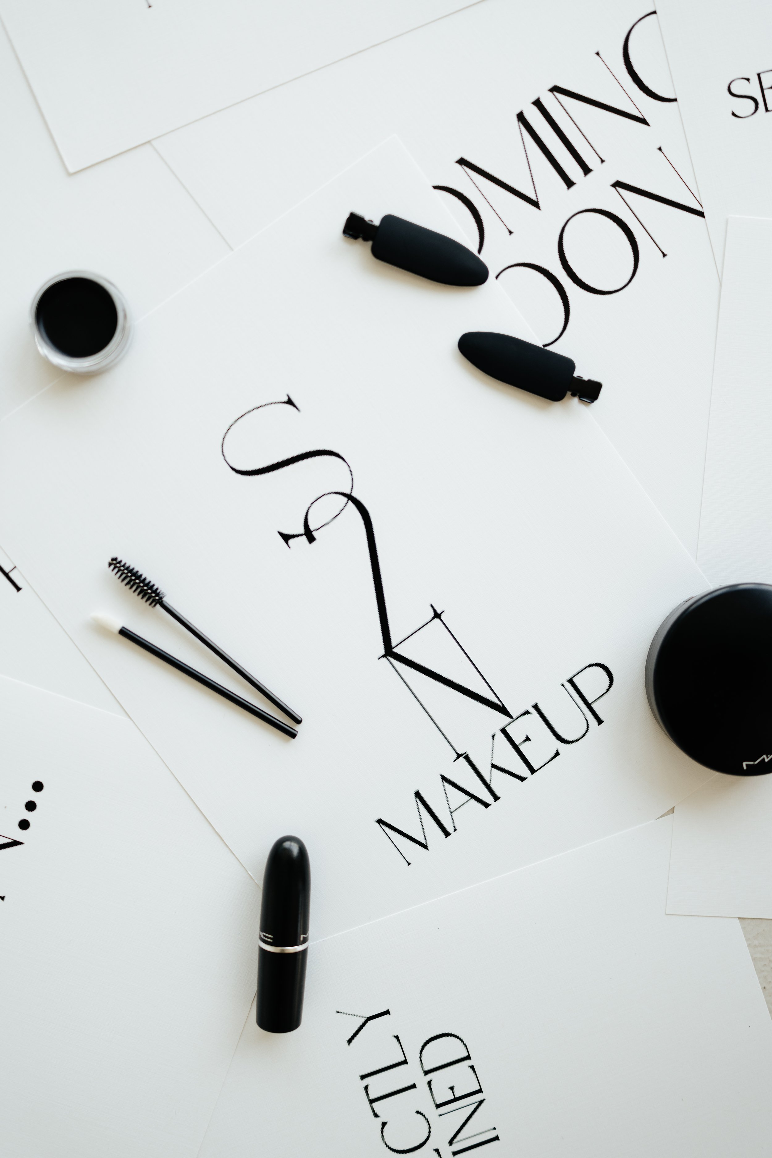Flat lay of makeup items and printed words on white paper, including brushes, black containers, and text such as "COMING SOON" and "MAKEUP."