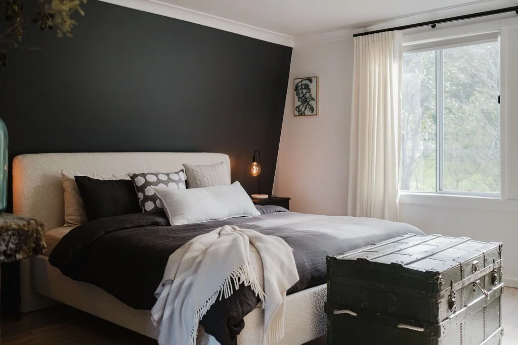 Modern bedroom with white and dark accent wall, bed with multiple pillows and blankets, vintage trunk at the foot of the bed, curtained window, and decorative wall art.