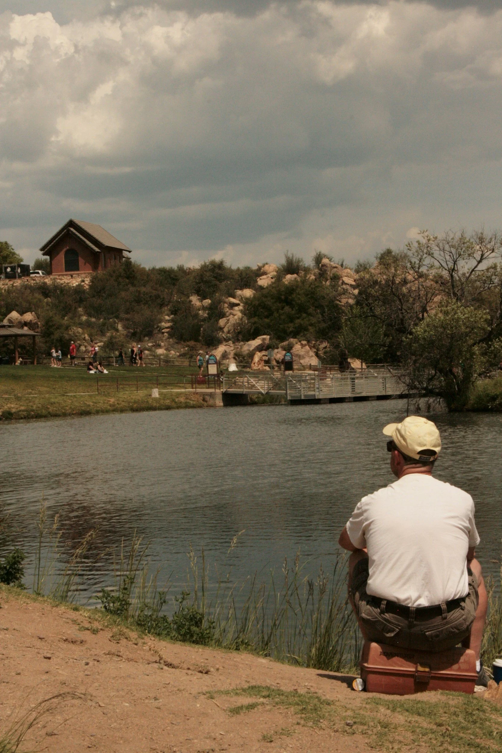 A man sitting on a cooler and looking at a pond in a park, with people walking and a building on a hill in the background.