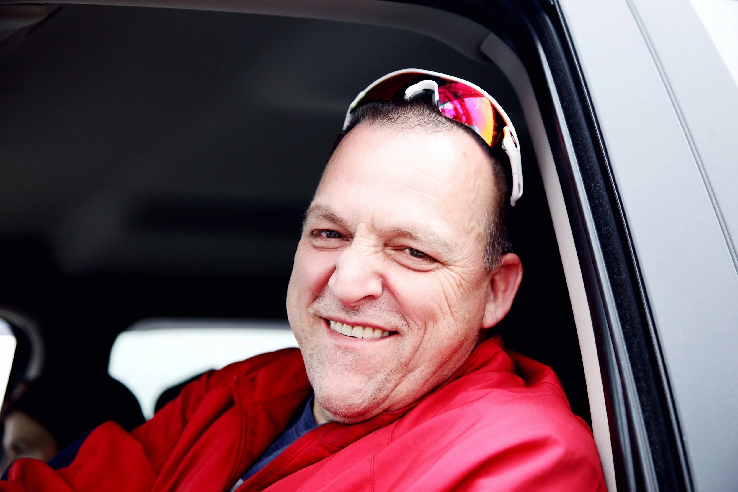 A smiling man with sunglasses resting on his head, wearing a red jacket, sitting in a vehicle.