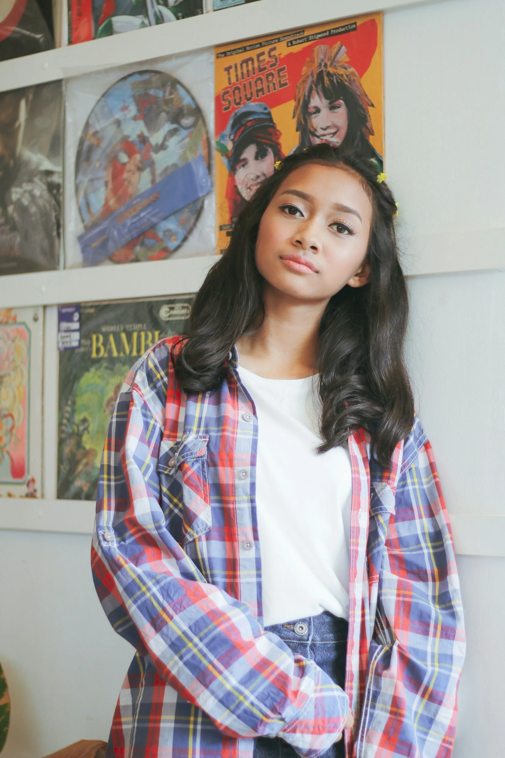 A young woman with long dark hair wearing a white t-shirt and a colorful plaid shirt standing in front of a wall decorated with posters, including "Times Square" and various comic book covers.