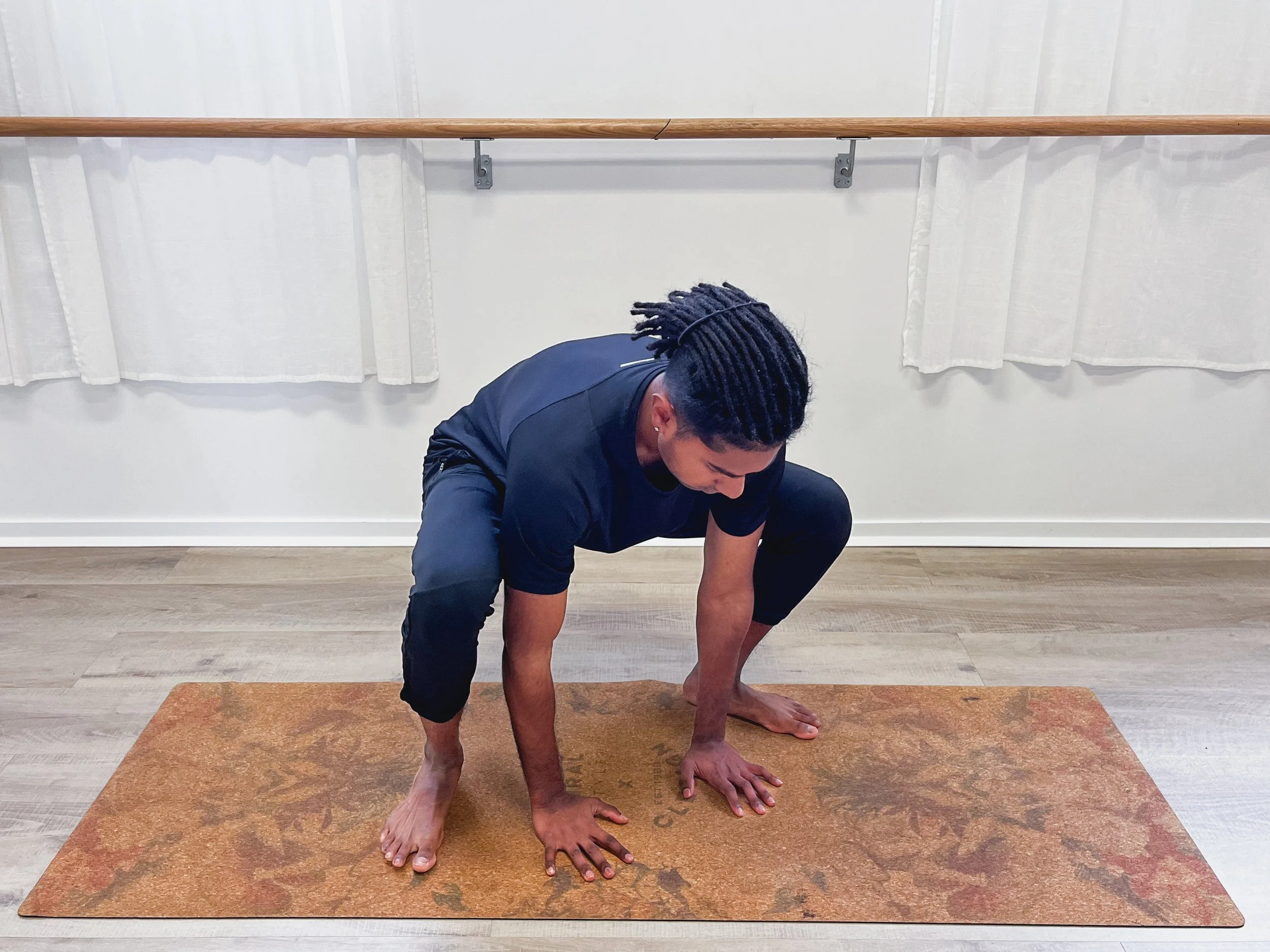 A person with dreadlocks in a black athletic outfit practicing yoga on an orange mat in a room with white curtains and a wooden ballet barre.