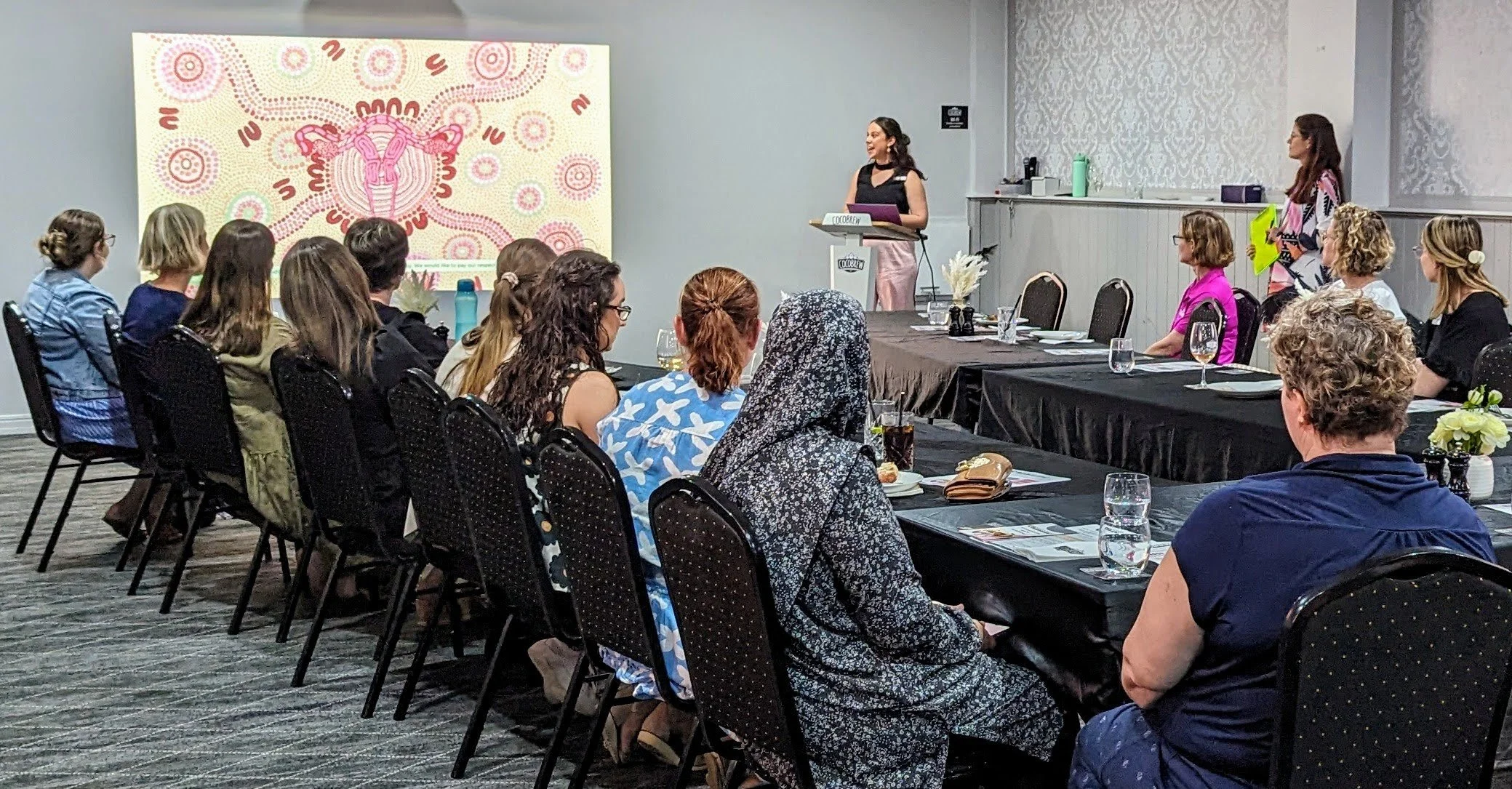 A conference room with women seated at black-covered tables, listening to a speaker at a podium, with a colorful artwork of the female reproductive system projected on a screen.