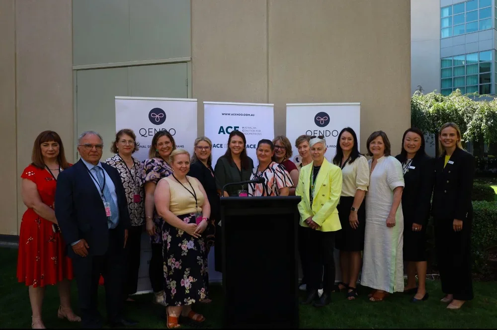 Group of people standing outdoors behind a black podium at a conference, with banners that read QENDO and ACE in the background.