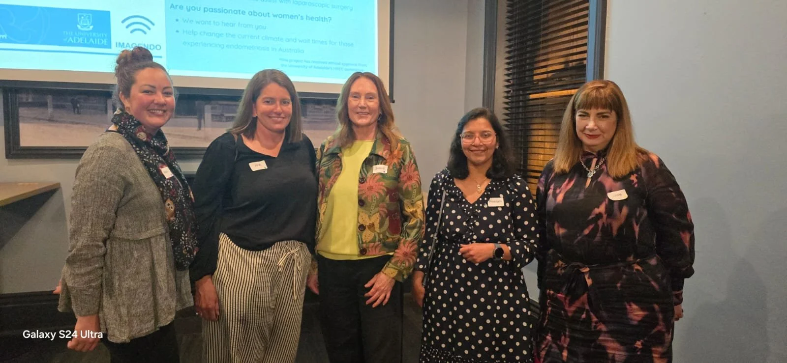 Group of five women standing together indoors, smiling, with a presentation screen behind them.