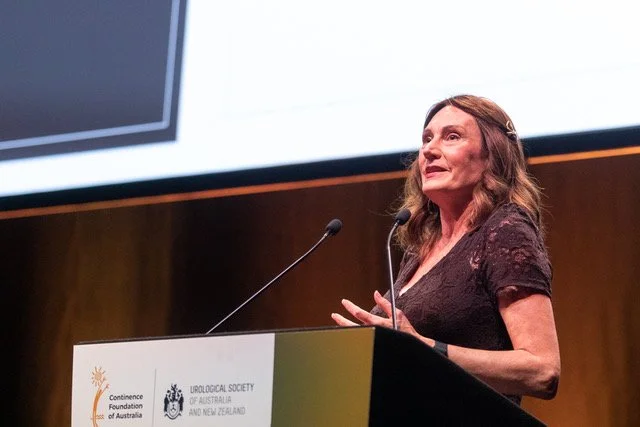 A/Prof Susan Evans speaking at a podium during a conference, with a large screen behind her displaying presentation slides.