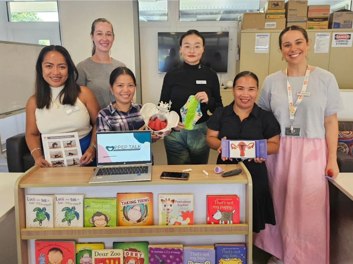 Group of six women standing behind a table with children's books, a laptop, and various items, in a room with boxes and cabinets in the background.
