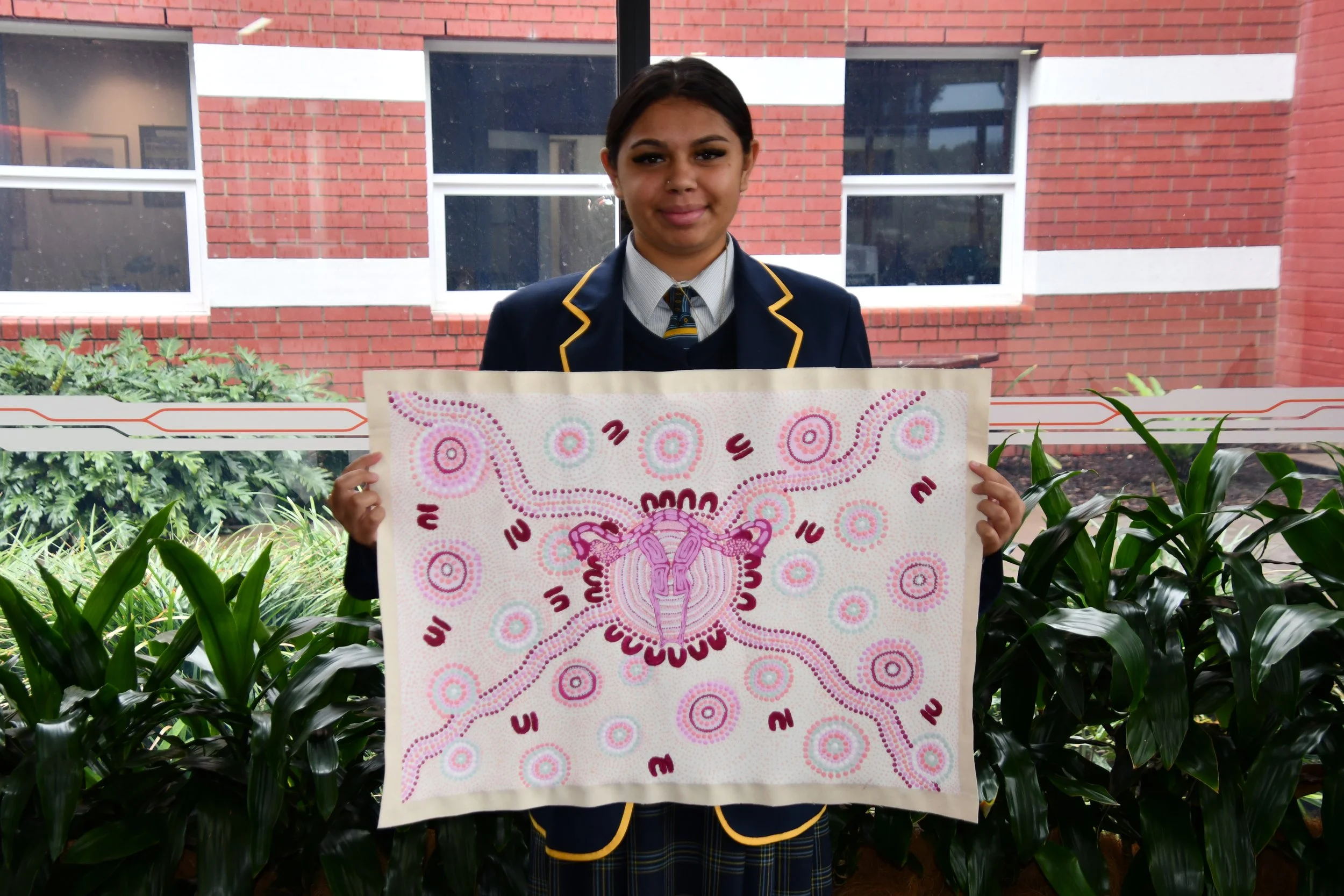 Young girl in school uniform holding a colorful Aboriginal-style artwork of a kangaroo's head, with patterns and dots, in front of a window with a brick building background.