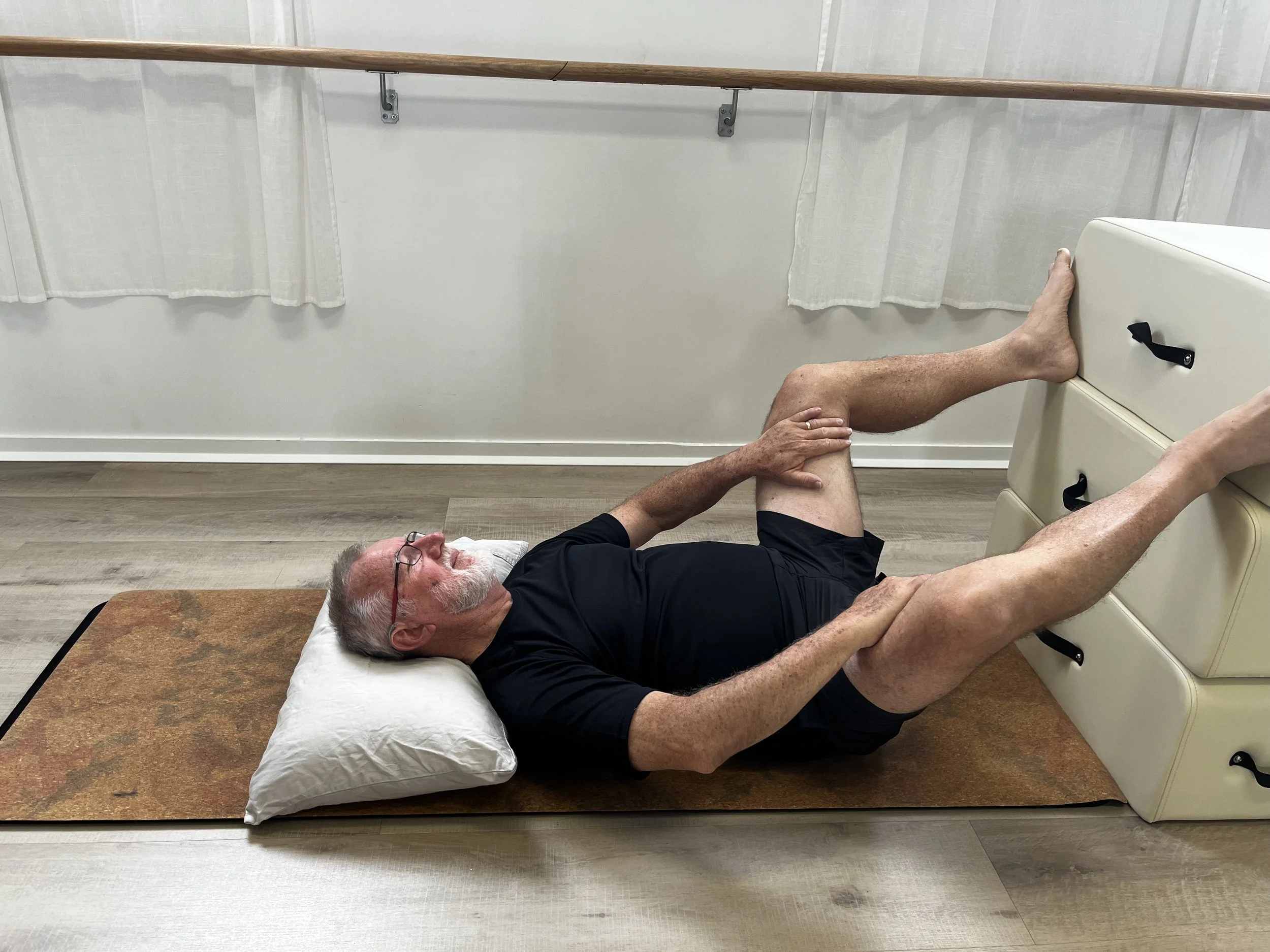 An elderly man lying on his back on a mat, with his head on a pillow, exercising his legs against a padded exercise box in a room with light-colored walls and hardwood flooring.