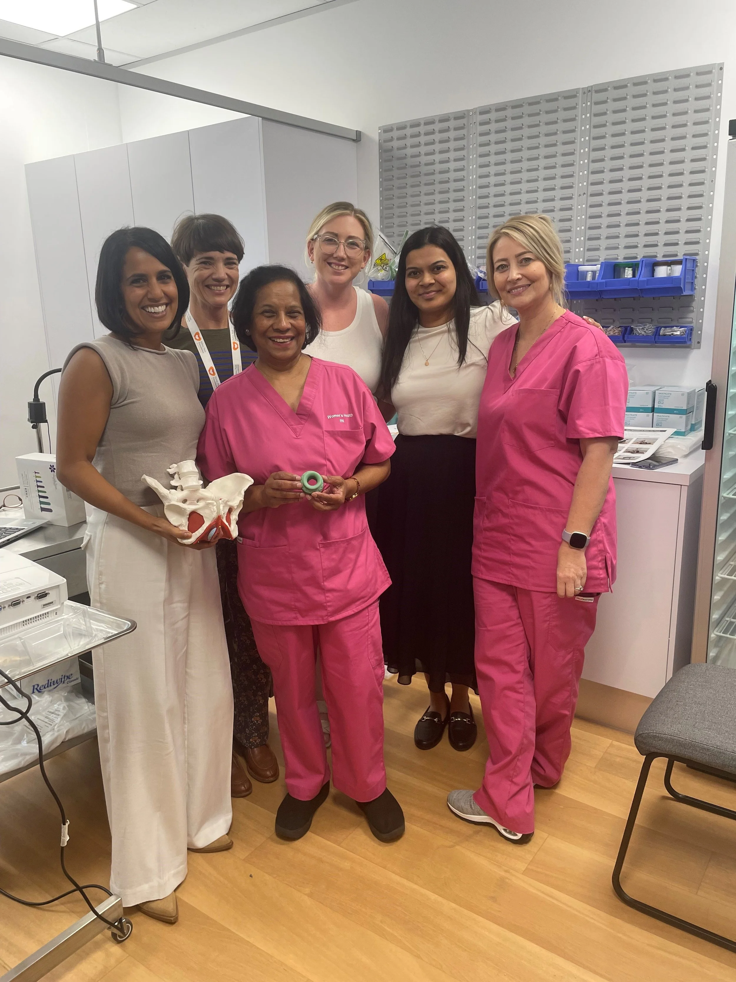 Evoca Women's Health. Group of five women in medical attire and casual clothing standing together in a clinical setting, smiling at the camera.