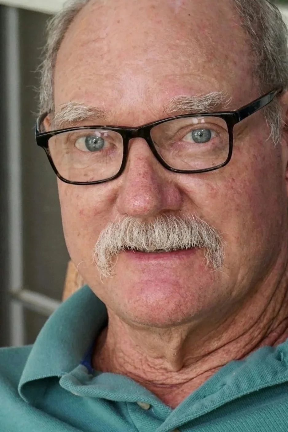 Close-up photo of an older man with gray hair, mustache, and glasses, wearing a teal polo shirt.