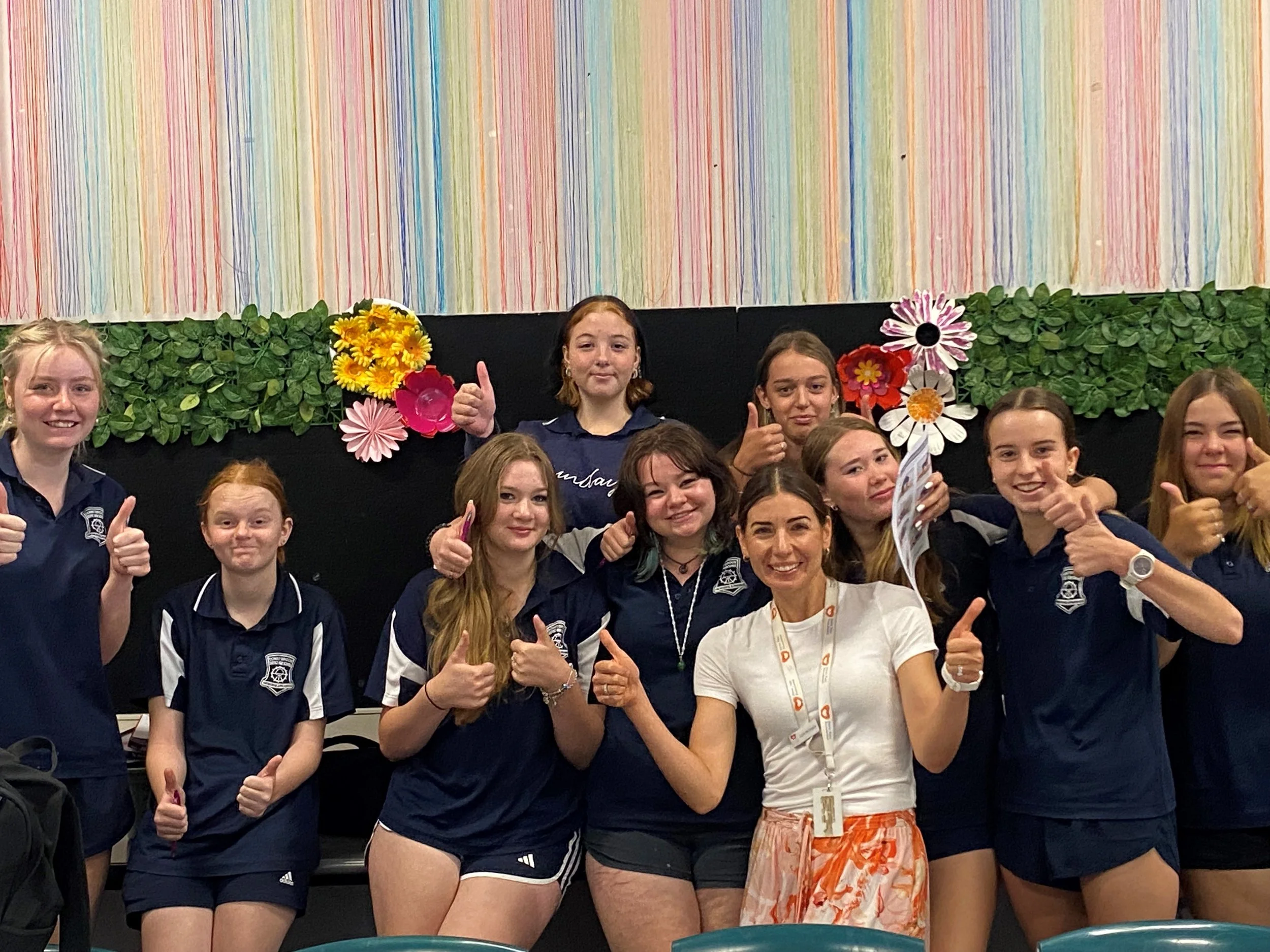 A group of ten young girls and one woman posing indoors with thumbs up, some holding flowers, in front of a colorful striped backdrop and green foliage decoration.