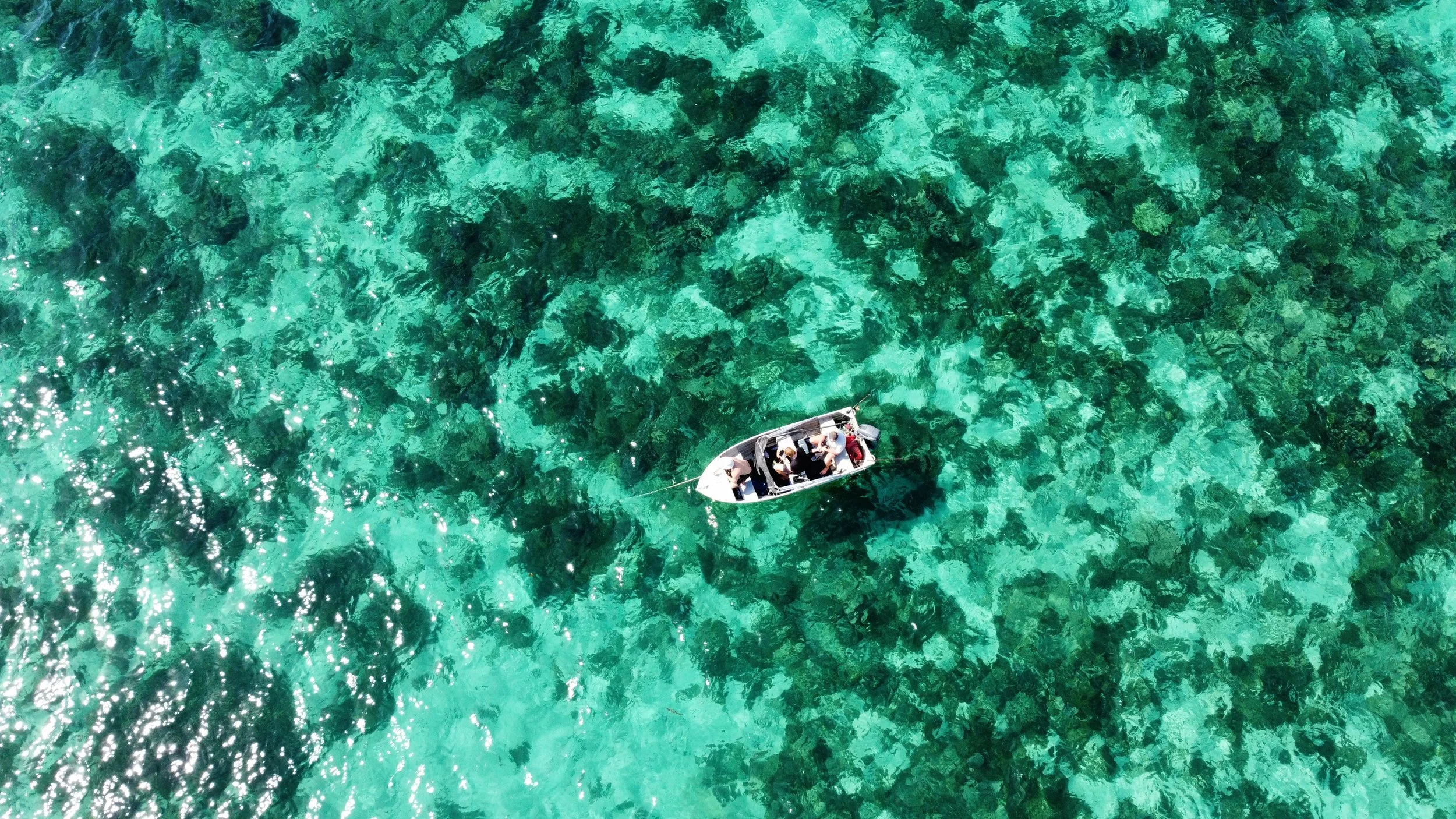 An aerial view of a small boat with people on board floating in clear turquoise water on the Ningaloo Reef, revealing the underwater rocks and coral beneath. With fishing and diving alongside