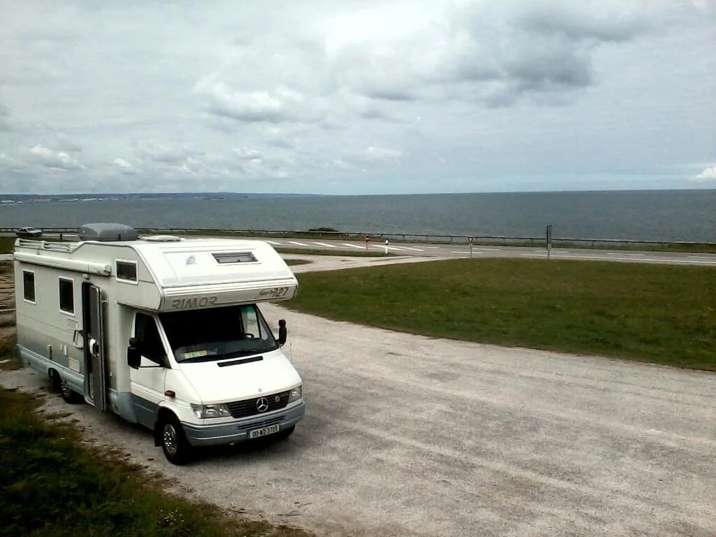A white camper van parked on a gravel area in Normandy, Francenear a grassy field overlooking a large body of water with cloudy skies overhead.