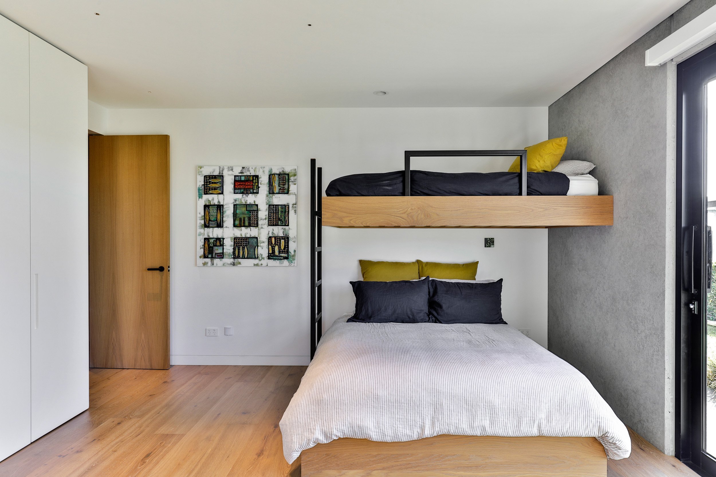 A modern bedroom featuring a double bunk bed with a wood frame, patterned artwork, and large windows, offering natural light.