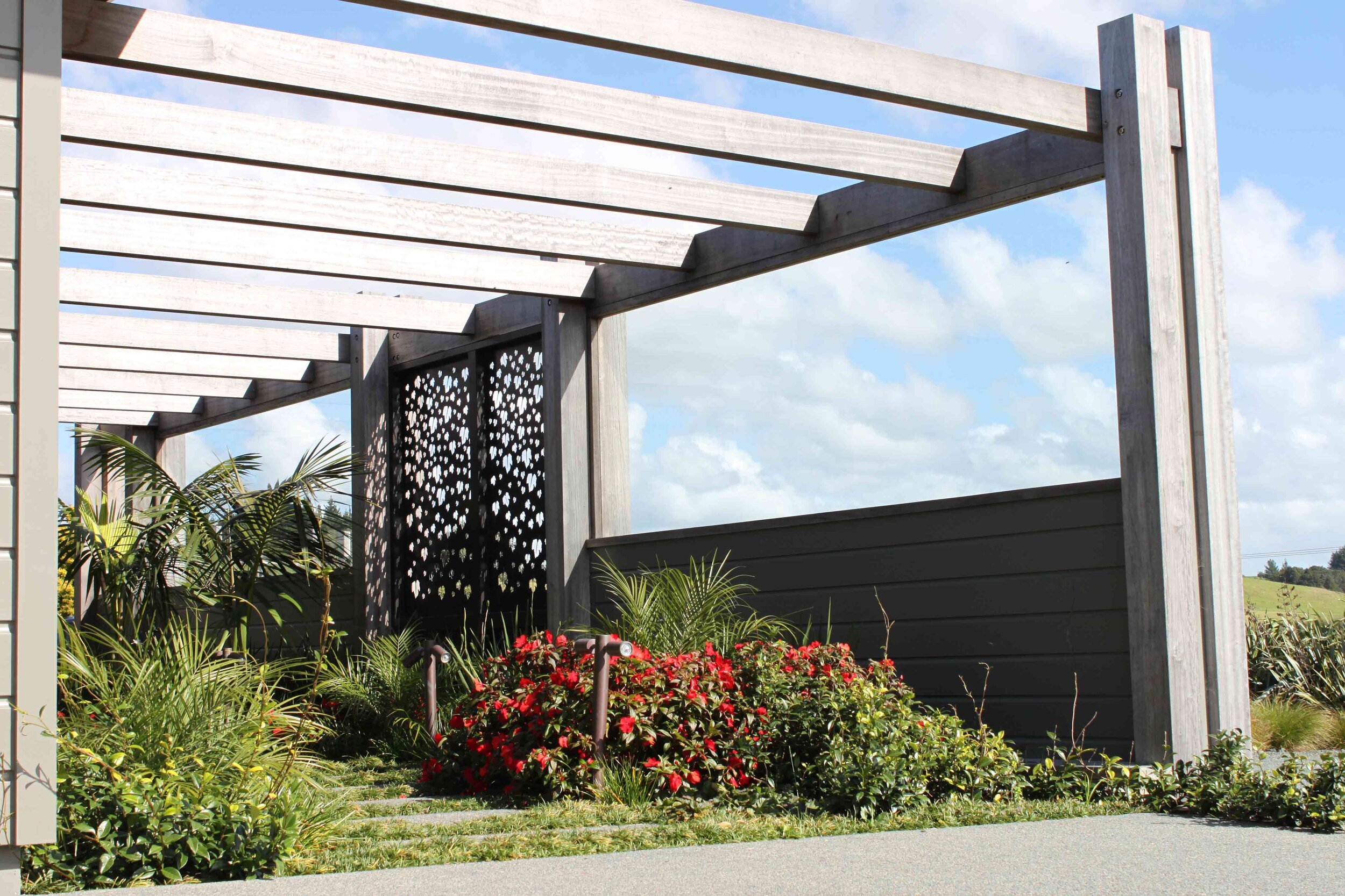 A modern pergola with a patterned screen surrounds vibrant plants and flowers, under a bright blue sky with fluffy clouds.