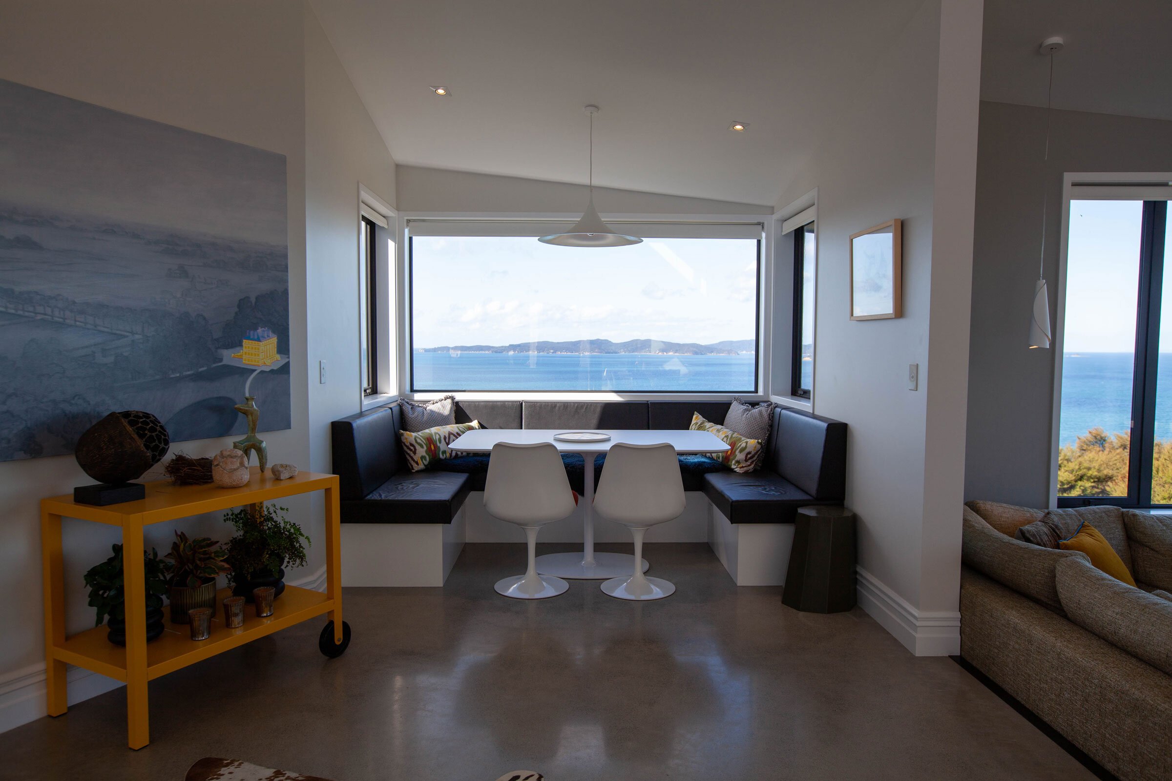 A bright, modern dining nook with a panoramic ocean view, featuring a white table, sleek chairs, and cozy cushions.