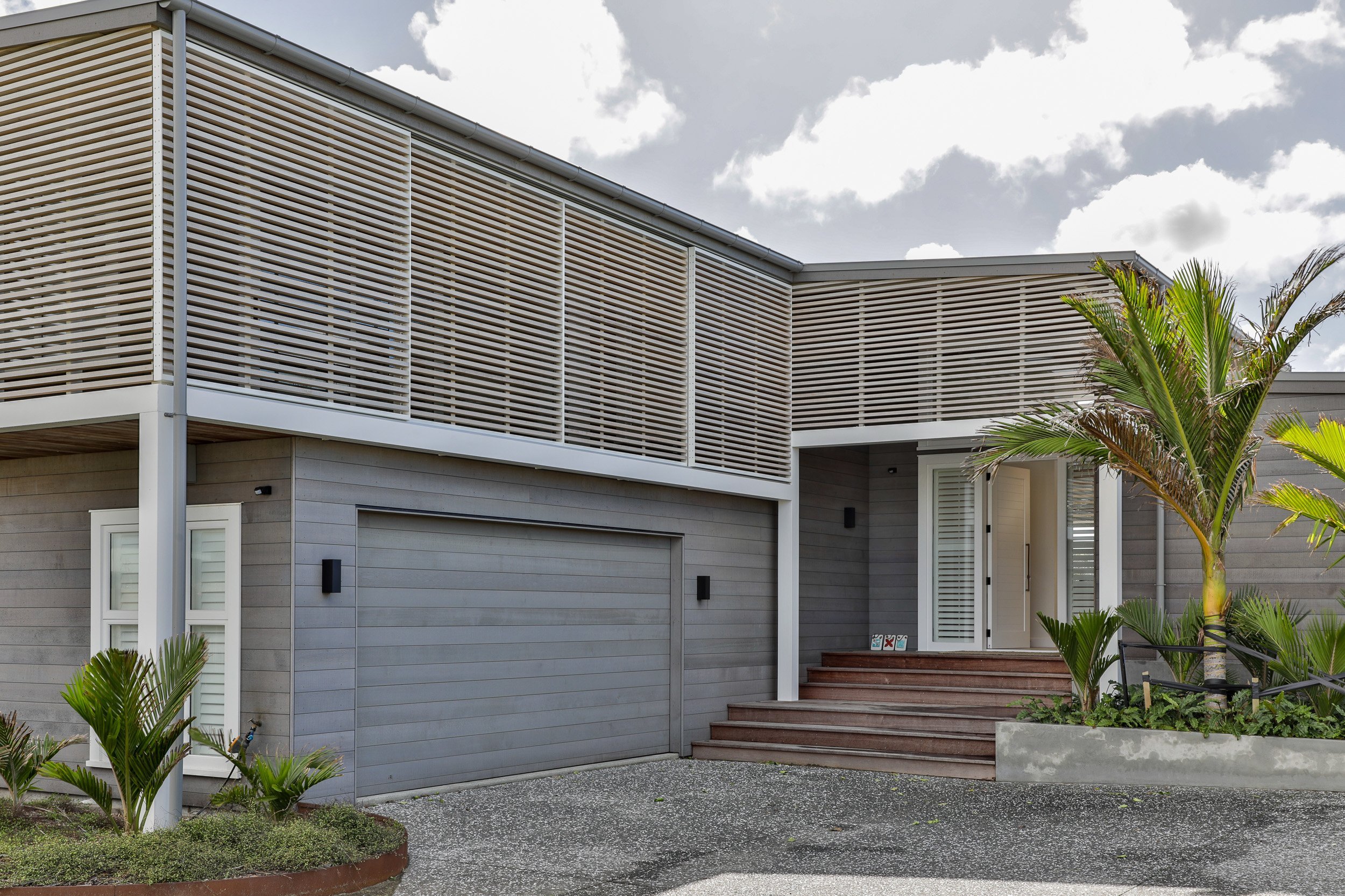 Modern house exterior with wooden slats, a driveway, steps leading to the entrance, and tropical plants in front. Cloudy sky above.