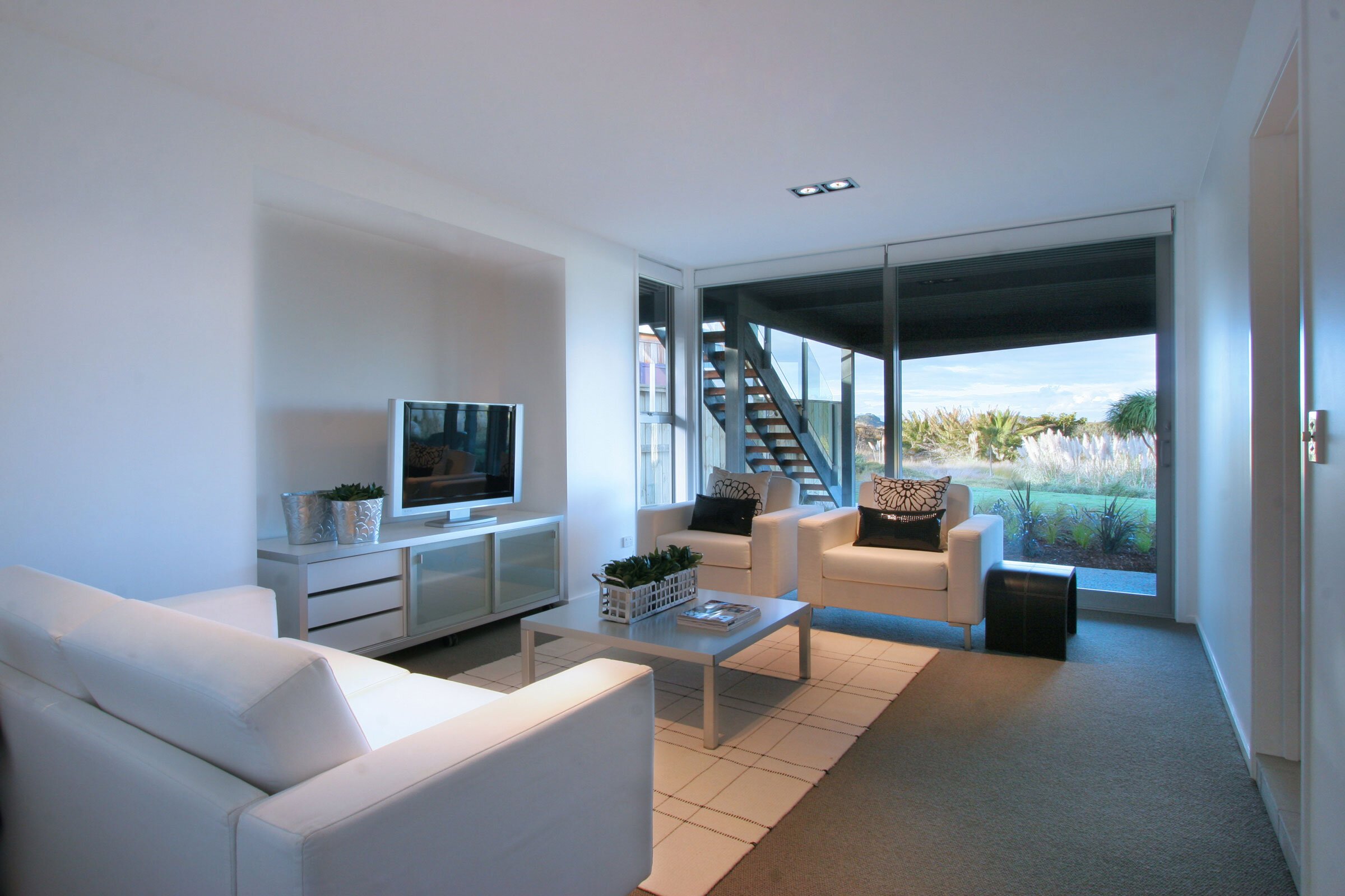 Bright and modern living room featuring white furniture, a TV, and large windows overlooking a landscaped outdoor area.